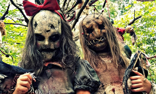 Two eerie dolls with cracked faces and long hair stand in a forest, surrounded by hanging creepy toys and branches.