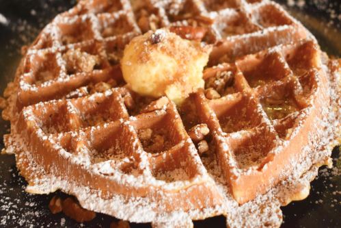 A close-up of a golden waffle topped with powdered sugar, a pat of butter, and chopped nuts.