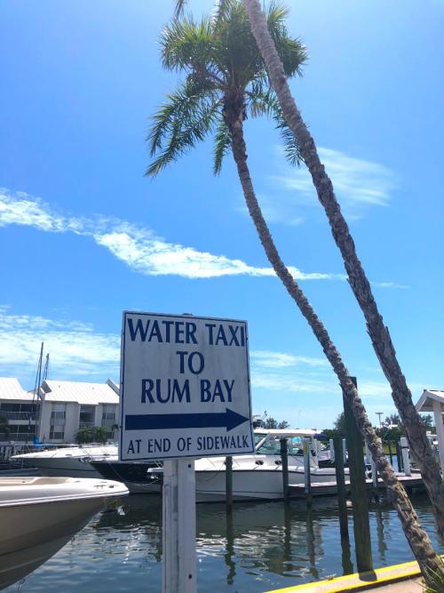 Sign for water taxi to Rum Bay at the end of the sidewalk, with a palm tree and boats in the background under a blue sky.