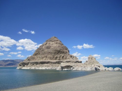 A rocky formation rises from a calm lake under a bright blue sky with scattered clouds.