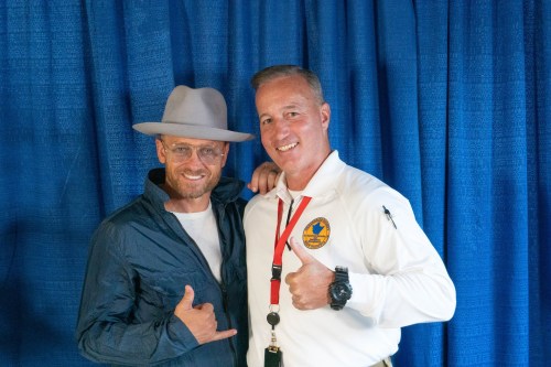 Two men pose together, one wearing a hat and jacket, the other in a white shirt with a badge, both smiling and giving thumbs up.