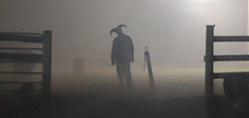 A shadowy figure with a horned mask stands in a foggy, dimly lit area near a fence.