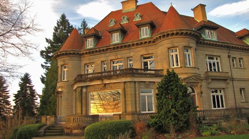 A grand, historic mansion with a tiled roof, large windows, and lush greenery surrounding it.