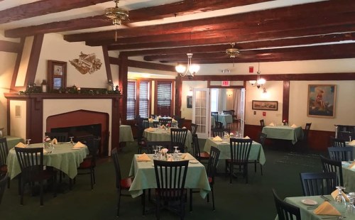 Cozy restaurant interior with green tablecloths, wooden beams, and a fireplace, featuring empty tables and warm lighting.