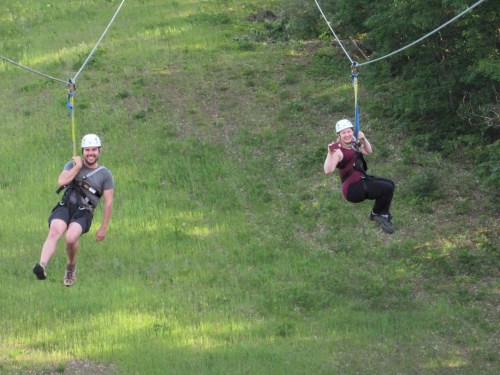 Two people ziplining through a green landscape, wearing helmets and harnesses, enjoying an outdoor adventure.