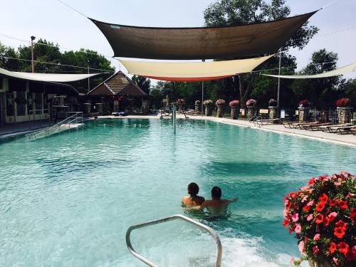 Two people relax in a swimming pool under shaded canopies, surrounded by flowers and lounge chairs.