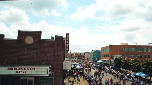 A bustling street scene with crowds, motorcycles, and shops under a partly cloudy sky.