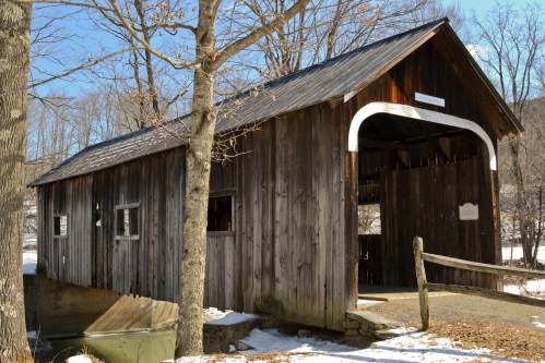 A rustic wooden covered bridge surrounded by trees and snow, with a clear blue sky in the background.