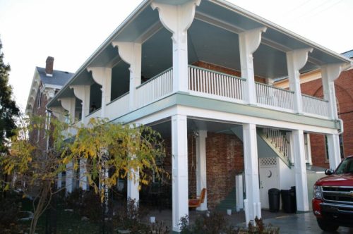 A two-story house with a large porch, white columns, and green accents, surrounded by trees and shrubs.