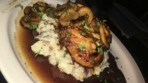 A plate of grilled chicken atop creamy mashed potatoes, garnished with sautéed mushrooms and fresh herbs.