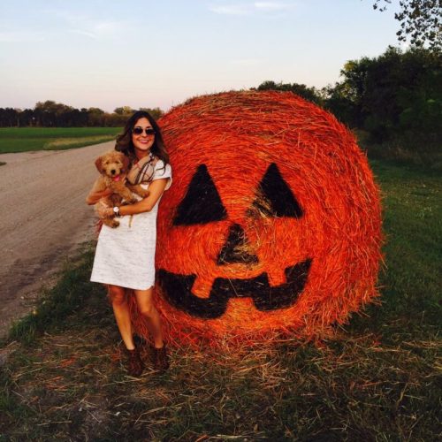 A woman in a white dress holds a small dog next to a large pumpkin-shaped hay bale in a rural setting.