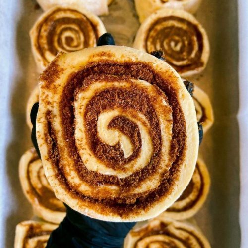 A close-up of a cinnamon roll with a spiral pattern, held by a gloved hand, surrounded by more rolls in the background.