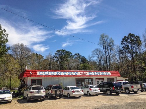 A roadside diner with a red roof, surrounded by trees and parked cars under a blue sky with wispy clouds.