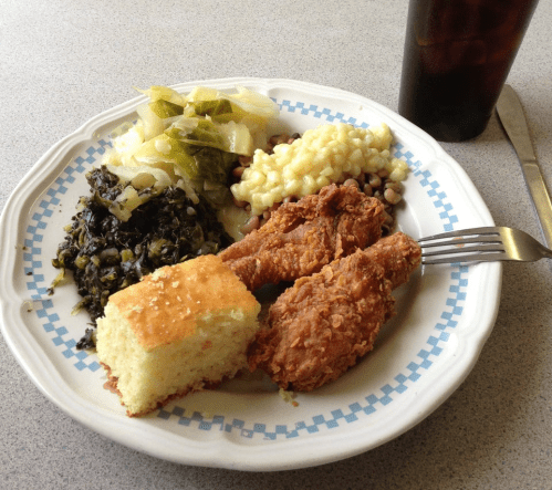 A plate of fried chicken, cornbread, collard greens, macaroni, and black-eyed peas, with a drink on the side.