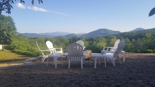 A serene outdoor setting with white chairs around a fire pit, surrounded by lush greenery and distant mountains.