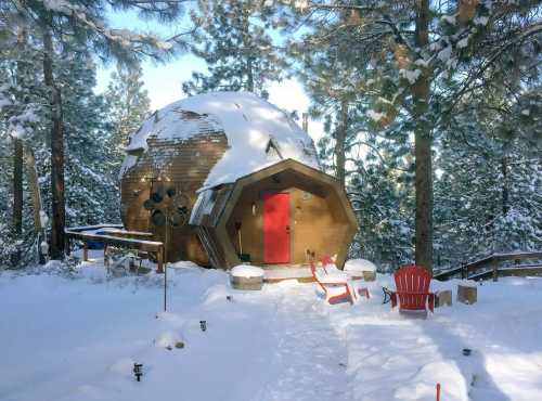 A unique, dome-shaped cabin surrounded by snow, featuring a red door and two red chairs in front. Pine trees in the background.