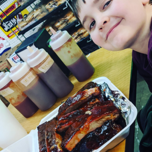 A smiling boy sits at a table with a plate of ribs and bottles of barbecue sauce in the background.