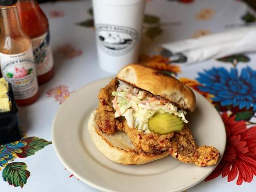 A fried chicken sandwich with pickles and coleslaw on a plate, accompanied by hot sauce bottles and a drink.
