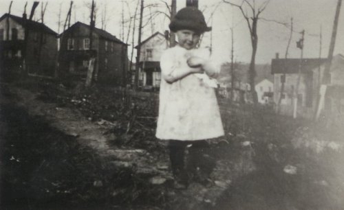 A young child in a hat holds a doll, standing on a path with houses and bare trees in the background.