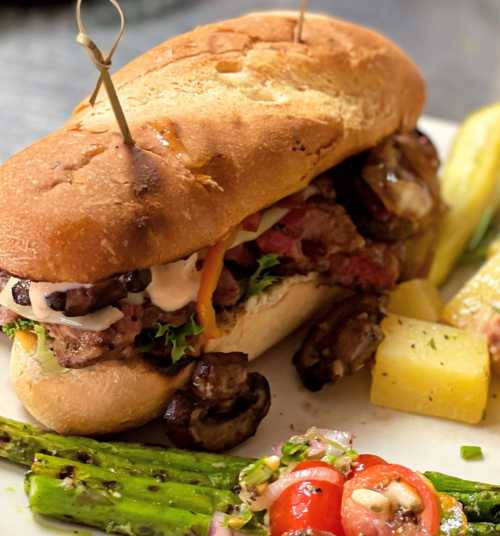 A hearty sandwich with beef, mushrooms, and toppings, served with asparagus and a side of diced pineapple and tomatoes.