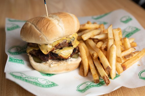 A juicy cheeseburger with pickles and sauce, served with a side of golden French fries on a paper-lined plate.