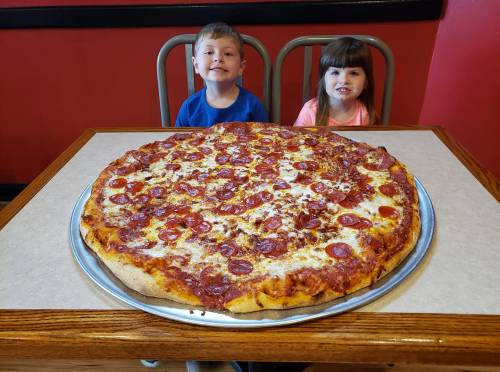 Two children sit at a table with a large pepperoni pizza in front of them, smiling at the camera.