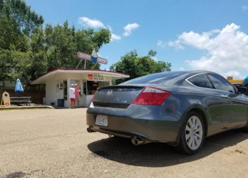 A gray car parked on a road near a small food stand with a blue sky and trees in the background.