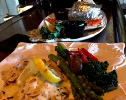 A plate of grilled fish with lemon, almonds, and vegetables, with another dish in the background.