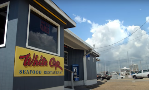 Exterior of White Cap Seafood Restaurant with a blue building, yellow sign, and cloudy sky in the background.