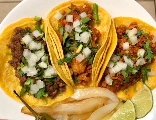 Three tacos on a plate, filled with meat, onions, and cilantro, accompanied by lime wedges and grilled onions.
