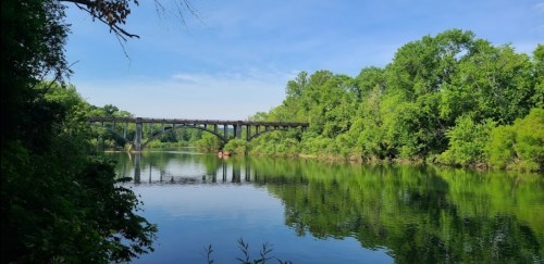 A serene river scene with lush green trees and a bridge reflecting in the calm water under a clear blue sky.