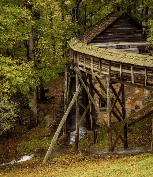A rustic wooden mill perched on stilts, surrounded by lush green trees and flowing water below.