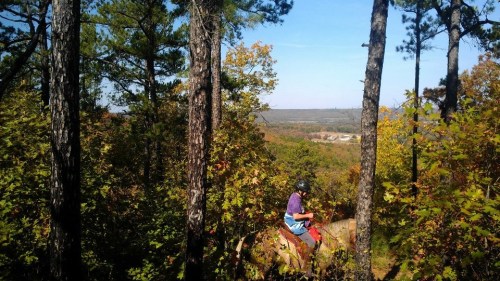 A person in a helmet sits on a rock, surrounded by trees and colorful autumn foliage, overlooking a valley.