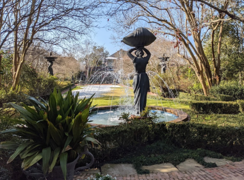 A garden scene featuring a fountain with a statue of a woman carrying a large bowl, surrounded by greenery and trees.