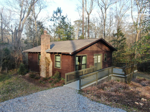 A rustic wooden cabin surrounded by trees, featuring a stone chimney and a front deck with a gravel pathway.