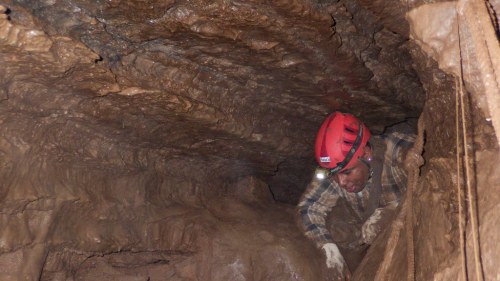 A person in a red helmet explores a rocky cave, using a headlamp to illuminate the dark surroundings.