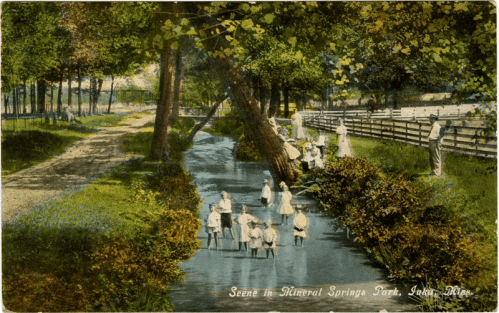 A vintage scene of children playing in a stream at Mineral Springs Park, surrounded by trees and a dirt path.