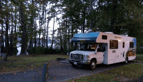 An RV parked in a wooded area near a calm lake, surrounded by trees at dusk.