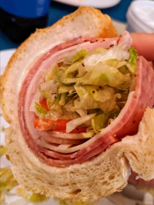 A close-up of a sandwich with layers of deli meats, lettuce, and tomato, held in a hand with a slice of bread.