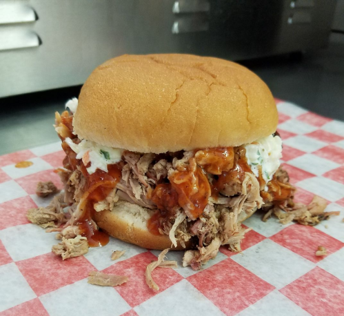 A pulled pork sandwich with barbecue sauce and coleslaw on a soft bun, served on a checkered paper.