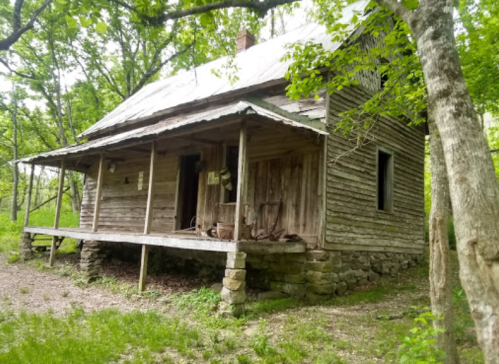 An old, weathered wooden house with a porch, surrounded by lush greenery and trees.