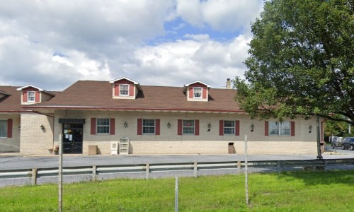 A single-story building with a brown roof, red shutters, and a grassy area in front under a partly cloudy sky.