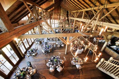 A spacious, rustic restaurant interior with wooden beams, round tables set for dining, and large windows letting in light.