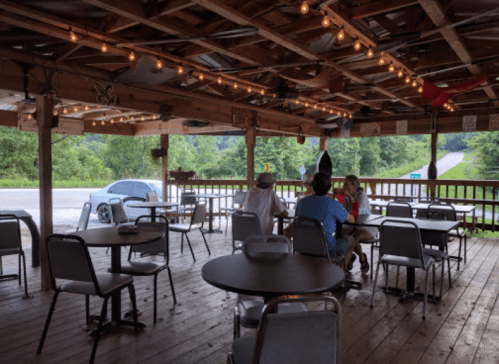 Outdoor dining area with string lights, empty tables, and a few patrons enjoying a meal. Green trees in the background.
