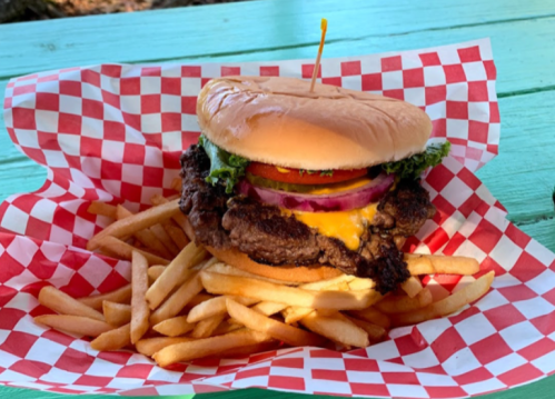 A large burger with lettuce, tomato, onion, and cheese, served with a side of golden fries on a red checkered paper.