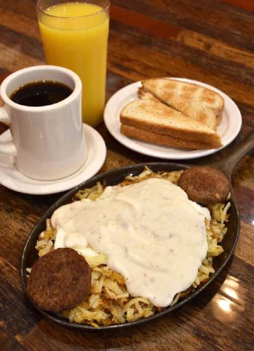 A skillet of hash browns topped with gravy, two sausage patties, toast, and a cup of coffee with orange juice.