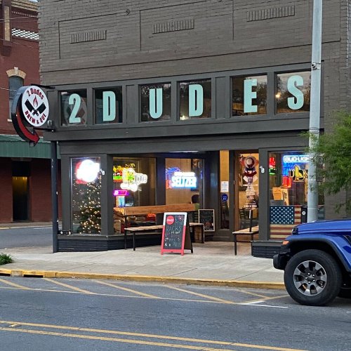 Exterior of a restaurant named "2 Dudes," featuring colorful signage and outdoor seating, with a parked blue vehicle nearby.