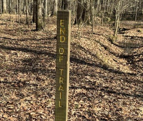 A wooden signpost reading "END OF TRAIL" in a forest with fallen leaves and trees in the background.