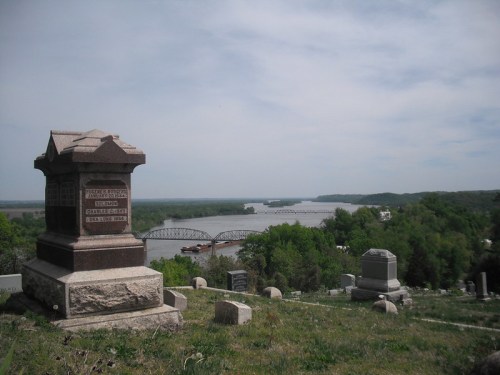 A scenic view from a cemetery, featuring a gravestone and a river with a bridge in the background.