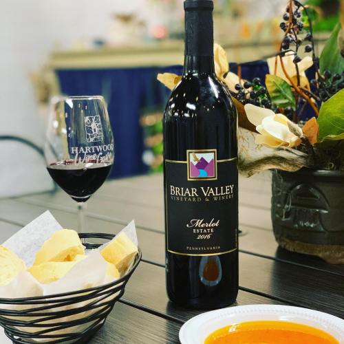 A bottle of Briar Valley Merlot wine next to a glass of red wine and a plate of snacks on a wooden table.